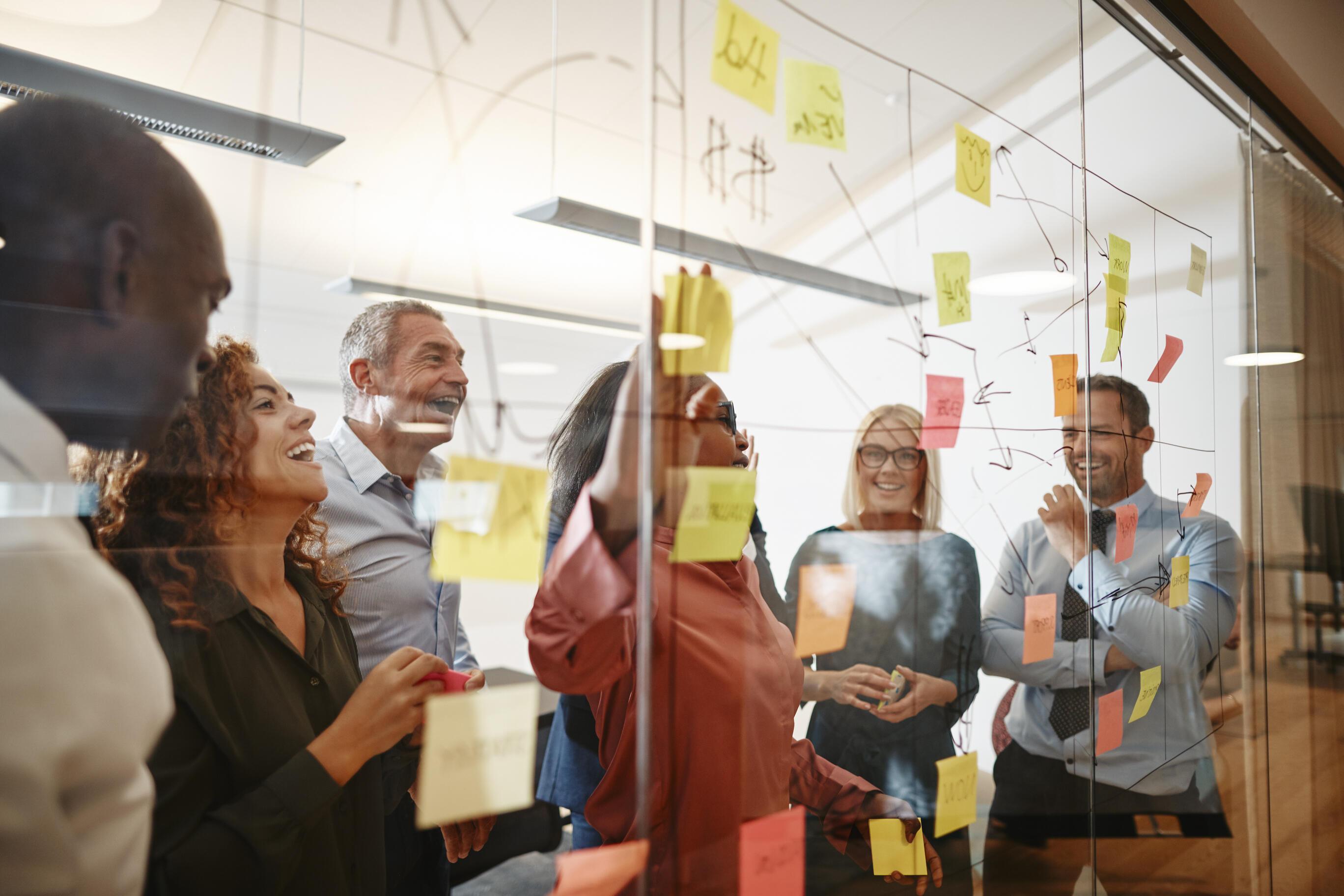 A diverse group of leaders laughing and collaborating around a glass wall covered with colorful sticky notes